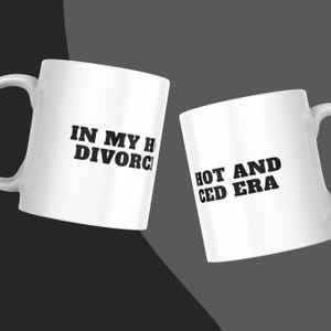 Könnte beinhalten: Zwei weiße Keramikbecher mit schwarzem Text. Der linke Becher zeigt die Worte IN MY HO DIVORC, während der rechte Becher die Worte HOT AND CED ERA zeigt. Beide Becher haben einen geschwungenen Henkel und eine glatte, glänzende Oberfläche.