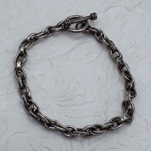 May include: A silver-toned metal chain bracelet featuring a series of interlocking oval links. The bracelet is secured with a toggle clasp, consisting of a circular ring and a T-shaped bar, both showing a polished finish with subtle dark oxidation.