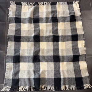 May include: A rectangular throw blanket featuring a large-scale plaid pattern in shades of black, charcoal gray, light gray, and off-white. The blanket has a textured, woven appearance and is finished with short, straight fringe along the top and bottom edges.