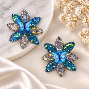 Könnte beinhalten: Zwei blumenförmige Broschen mit abwechselnden Blütenblättern aus irisierendem Silber und tiefblauem Glitzer. Jede Brosche hat in der Mitte eine kleine hellblaue Schmetterlingsfigur. Die Blütenblätter sind sternförmig mit strukturierter Oberfläche angeordnet.
