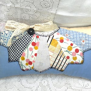 May include: A rectangular light blue pillow featuring a central fan-shaped patchwork applique made of various patterned fabrics including gingham, floral, and striped prints. The applique is accented with a cream lace bow and a small black button.