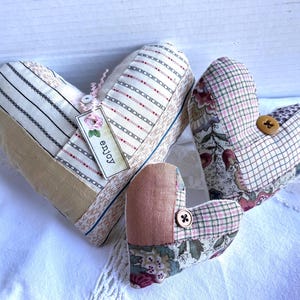 May include: Three heart-shaped fabric ornaments in varying sizes. The largest features striped and solid beige panels with a tag reading "enjoy". The two smaller hearts are made of floral and plaid patchwork fabric, each accented with a single brown button.
