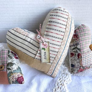 May include: Three heart-shaped fabric ornaments in varying sizes. The largest heart features striped fabric and a tag with a rose illustration and the word enjoy. The smaller hearts feature floral and plaid patterns with brown buttons attached.