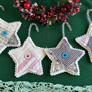 May include: Four handmade star-shaped Christmas ornaments. Each star is made of fabric patches in various patterns and colors, including pink stripes, gingham, and floral prints. Each star has a button in the center and a silver wire loop for hanging.