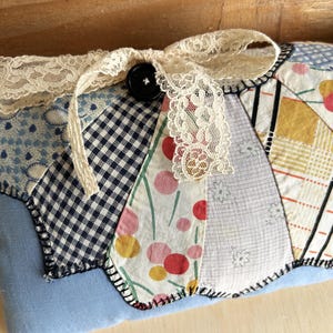 May include: A handmade patchwork pillow with a light blue base and a variety of patterned fabric sections. The sections include gingham, floral, and patterned fabrics. A cream-colored lace bow and a black button adorn the top, adding a vintage touch.