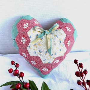 May include: A heart-shaped fabric cushion constructed from patchwork hexagons. The center features a cream floral print, surrounded by red hexagons with white flower motifs and a light green border. A small fabric bow is tied at the top.