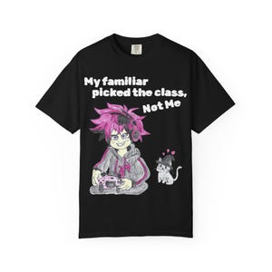 Może przedstawiać: Czarny t-shirt z grafiką przedstawiającą postać z różowymi włosami w słuchawkach, trzymającą różowy kontroler do gier. Obok postaci znajduje się mały biały kot w kapeluszu czarownicy. Tekst głosi: My familiar picked the class, Not Me.