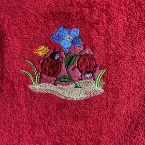 Könnte beinhalten: Ein besticktes rotes Handtuch mit einer blauen Blume und zwei roten Marienkäfern. Die Marienkäfer sind von grünen Blättern und einem gelben Schmetterling umgeben.