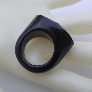 May include: A black ring with a geometric design. The ring has a thick band and a faceted, rectangular top. The ring is made of a glossy, dark material.