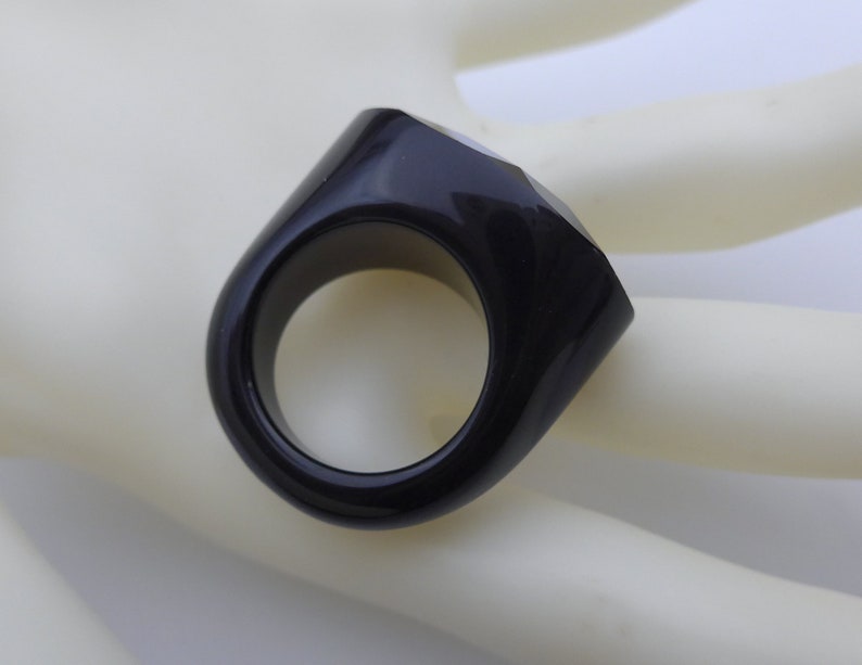 May include: A black ring with a geometric design. The ring has a thick band and a faceted, rectangular top. The ring is made of a glossy, dark material.