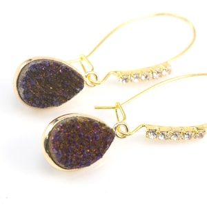 Pode incluir: Um par de brincos de argola dourados com uma pedra de ágata roxa em forma de lágrima e uma fileira de strass transparentes.