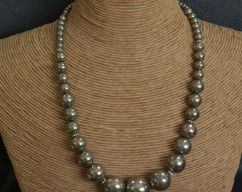 May include: A necklace made of dark gray round beads with a larger, round bead in the center. The necklace is displayed on a brown, textured mannequin.