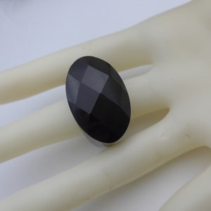May include: A large, oval-shaped black gemstone with a faceted surface. The gemstone is set on a ring, creating a bold and elegant statement piece. The dark color and geometric cuts give it a modern look.