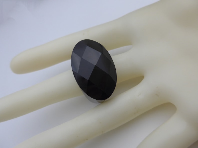 May include: A large, oval-shaped black gemstone with a faceted surface. The gemstone is set on a ring, creating a bold and elegant statement piece. The dark color and geometric cuts give it a modern look.