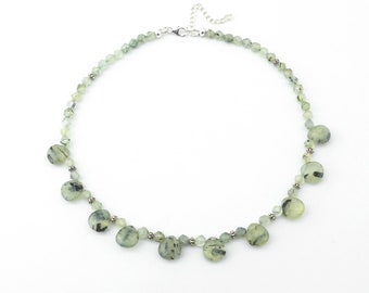 May include: A delicate necklace featuring a strand of small, faceted green beads and larger, rounded green stones with black inclusions. The necklace has a silver-toned clasp and connecting chain, creating a soft, natural aesthetic.