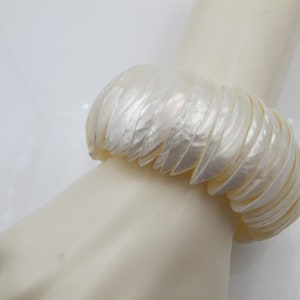 May include: A wide, off-white bracelet made of layered, shell-like segments. The bracelet is worn on a mannequin hand, showcasing its textured, iridescent surface. The design suggests a natural, organic theme.