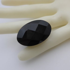 May include: A faceted, oval-shaped black gemstone. The gem has a polished surface with multiple flat planes that reflect light. The stone is set against a neutral background.