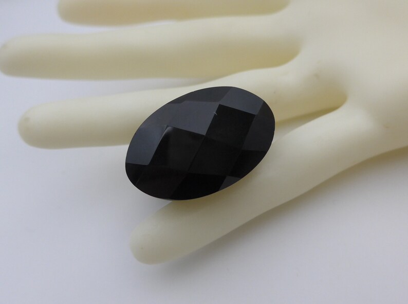 May include: A faceted, oval-shaped black gemstone. The gem has a polished surface with multiple flat planes that reflect light. The stone is set against a neutral background.