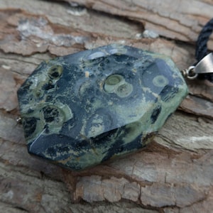 May include: A green and black stone pendant with a unique pattern, hanging from a black cord. The stone has a polished, faceted surface.