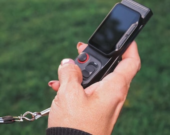 May include: A black handheld electronic device with a rectangular screen, a red circular joystick, and two black buttons. The device features a metallic side frame and is attached to a thin black lanyard with a silver metal clasp.