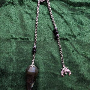 May include: A pendulum featuring a faceted, dark, iridescent gemstone point attached to a silver-toned metal chain. The chain is accented with two small, dark, spherical beads on each side and terminates with a small, silver-toned metal bat charm.