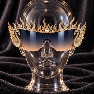 Pode incluir: Óculos de sol dourados com uma armação superior em forma de chama adornada com cristais transparentes. Cada lado da armação apresenta um motivo de cisne com um olho de cristal azul. As lentes são de estilo máscara com um degradê de cor.