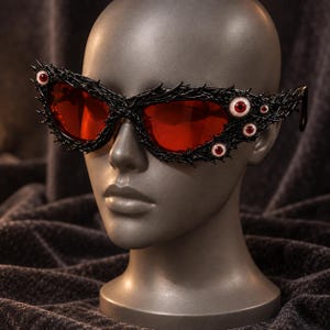 May include: Black sunglasses featuring a textured, thorn-like frame adorned with multiple realistic red-irised eyeballs. The lenses are a deep, translucent red, and the frame extends outwards with sharp, pointed details surrounding the eyes.
