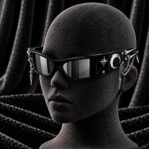 Op de afbeelding: Zwarte zonnebril met donker reflecterende glazen en een dik montuur. De pootjes zijn versierd met zilverkleurige metalen accenten, waaronder een ster, een halve maan, stekels en bungelende kettingbedels aan de zijkant van het montuur.