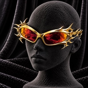Könnte beinhalten: Goldfarbene Sonnenbrille mit einem komplexen, dornenartigen Rahmendesign. Die Gläser haben einen leuchtenden, reflektierenden orange-roten Farbton. Der Rahmen läuft an den Bügeln und am Nasensteg in spitze, gezackte Kanten aus.