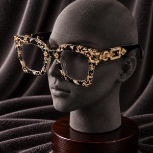 May include: Square-framed spectacles featuring a black and white marbled pattern on the rims, accented with intricate gold-tone filigree overlays. The temples are connected by a gold-tone chain-link hinge design, transitioning into solid black arms.