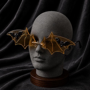 Op de afbeelding: Randloze zonnebril met amberkleurige glazen in de vorm van vleermuisvleugels. Elk glas is versierd met een kleine, zwarte, driedimensionale vleermuisfiguur aan de buitenrand. De bril heeft dunne metalen pootjes en een goudkleurige neusbrug.