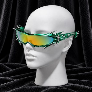 Op de afbeelding: Wraparound-zonnebril met een metallic groen montuur met scherpe, doornachtige punten langs de randen. De glazen hebben een reflecterende, oranje en gele kleurverloop. Een klein zilveren kruis-embleem is bevestigd op het tempelgedeelte.
