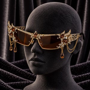 May include: Gold-tone sunglasses featuring brown lenses, embellished with clear crystals and amber-coloured teardrop gems. The frame includes intricate metal filigree, decorative wing-like side accents, and delicate gold chains hanging from the edges.
