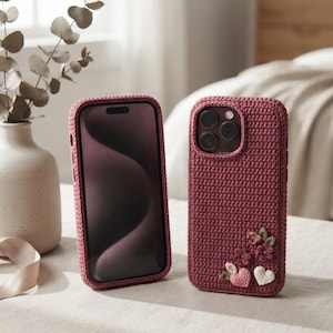 Può includere: Due custodie per smartphone con una texture all'uncinetto color rosa scuro. Una custodia mostra lo schermo anteriore, mentre l'altra mostra il retro con piccole decorazioni all'uncinetto a forma di fiori e cuori nei toni del rosa, verde e bianco.