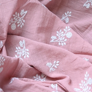 May include: A dusty rose pink fabric featuring a repeating white embroidered floral pattern. The textile has a visible woven texture with soft folds and gathers that create depth across the surface of the material.