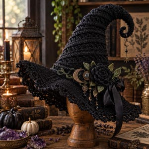 May include: A black crocheted witch hat featuring a pointed, curved top with a dangling moon and star charm. The hat is adorned with a large black crocheted rose, green crocheted leaves, a black ribbon, and metallic charms including a crescent moon.