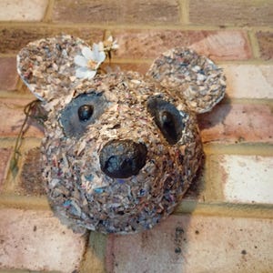 Könnte beinhalten: Eine bärenförmige Wanddekoration aus strukturiertem, mehrfarbigem Papierschnipsel-Material. Der Bär hat zwei abgerundete Ohren, eine dunkel-schwarze Nase und zwei dunkel-schwarze Augenhöhlen. Eine kleine weiße Kunstblume ist in der Nähe des linken Ohrs befestigt.