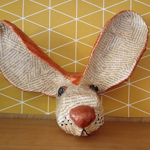 Könnte beinhalten: Eine Wandskulptur eines Kaninchenkopfes aus Decoupage-Papier mit gedrucktem Text. Das Kaninchen hat große, aufrechte Ohren mit kupferfarbenen Rändern, eine markante kupferfarbene Nase und kleine, dunkle Augen.