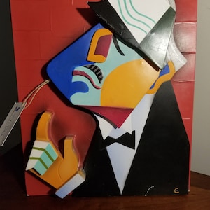 May include: A colorful abstract wall sculpture of a person wearing a white and green hat, a blue and yellow shirt, and a black tuxedo. The sculpture is made of wood and painted in bright colors.