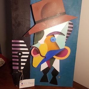 May include: Abstract art piece featuring a stylized face with a brown hat, blue eye, and colorful geometric shapes. The artwork is mounted on a textured blue background with a black border. A small tag reads "T.L. Design."