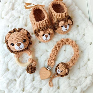 Könnte beinhalten: Ein Set aus beigen gehäkelten Babyschuhen, einer Löwen-Rassel mit Holzring und einem Schnullerhalter mit geflochtenem Band, herzförmigem Holzclip und einem kleinen gehäkelten Löwengesicht aus hellbraunem Garn.
