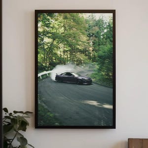 May include: A framed wall art print featuring a dark purple sports car drifting around a sharp curve on a paved mountain road. The vehicle is surrounded by a thick cloud of white tire smoke, set against a backdrop of dense green forest trees.