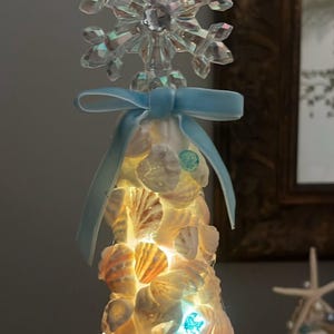 May include: A conical decorative tree covered in various white and tan seashells, illuminated by internal warm white lights. The top features a clear, faceted crystal snowflake ornament tied with a light blue satin ribbon bow.