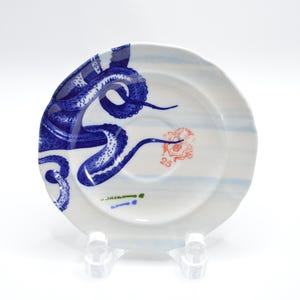 May include: A white ceramic saucer featuring a vibrant blue octopus tentacle design wrapping around the left side. The center includes a small orange crest with the number 25, faint light blue horizontal stripes, and small green text.