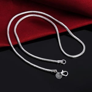 Op de afbeelding: Een zilverkleurige slangenketting met een gladde, flexibele textuur. De ketting is voorzien van een karabijnsluiting en een klein, rond metalen label met het getal 925, wat duidt op een samenstelling van sterling zilver voor dit sieraad.