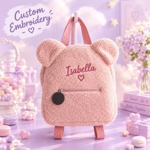 Op de afbeelding: Een roze rugzak in de vorm van een teddybeer, gemaakt van zacht, pluizig sherpamateriaal. Op de voorkant staat de naam Isabella geborduurd boven een klein hartje. De tas heeft een voorvak met rits en een ronde zwarte ritstrekker.