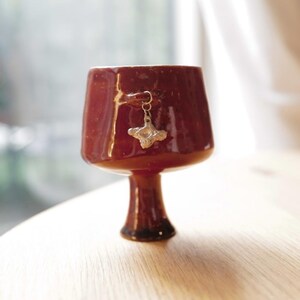 May include: A glossy, deep reddish-brown ceramic goblet with a flared rim and a pedestal base. A small, metallic, cross-shaped charm hangs from a thin metal ring attached to the side of the cup, resting against the smooth, reflective surface.