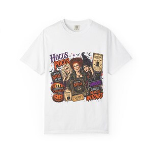 Może przedstawiać: Biały t-shirt z kolorowym nadrukiem graficznym przedstawiającym trzy czarownice, księgę zaklęć, dynie i czarnego kota. Tekst zawiera Hocus Pocus, I Put A Spell On You, Come Little Children, Book Of Spells, Black Flame Candle, Amuck oraz Magic Witches.