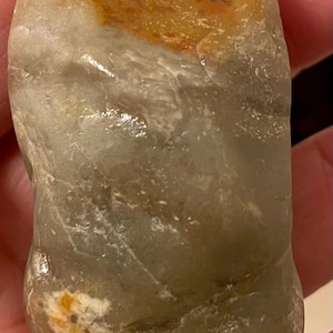 May include: A smooth, polished, translucent gray stone with a rounded, irregular shape. The surface features a prominent patch of warm orange-brown coloration near the top and a smaller, textured white and orange inclusion near the base.