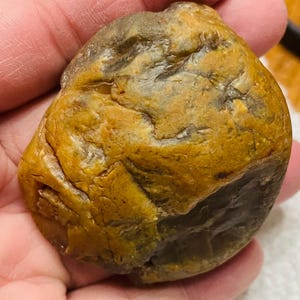 May include: A smooth, rounded stone featuring a mottled surface with deep ochre, mustard yellow, and dark brown tones. The rock has an irregular, textured appearance with visible fissures and indentations across its polished, organic form.